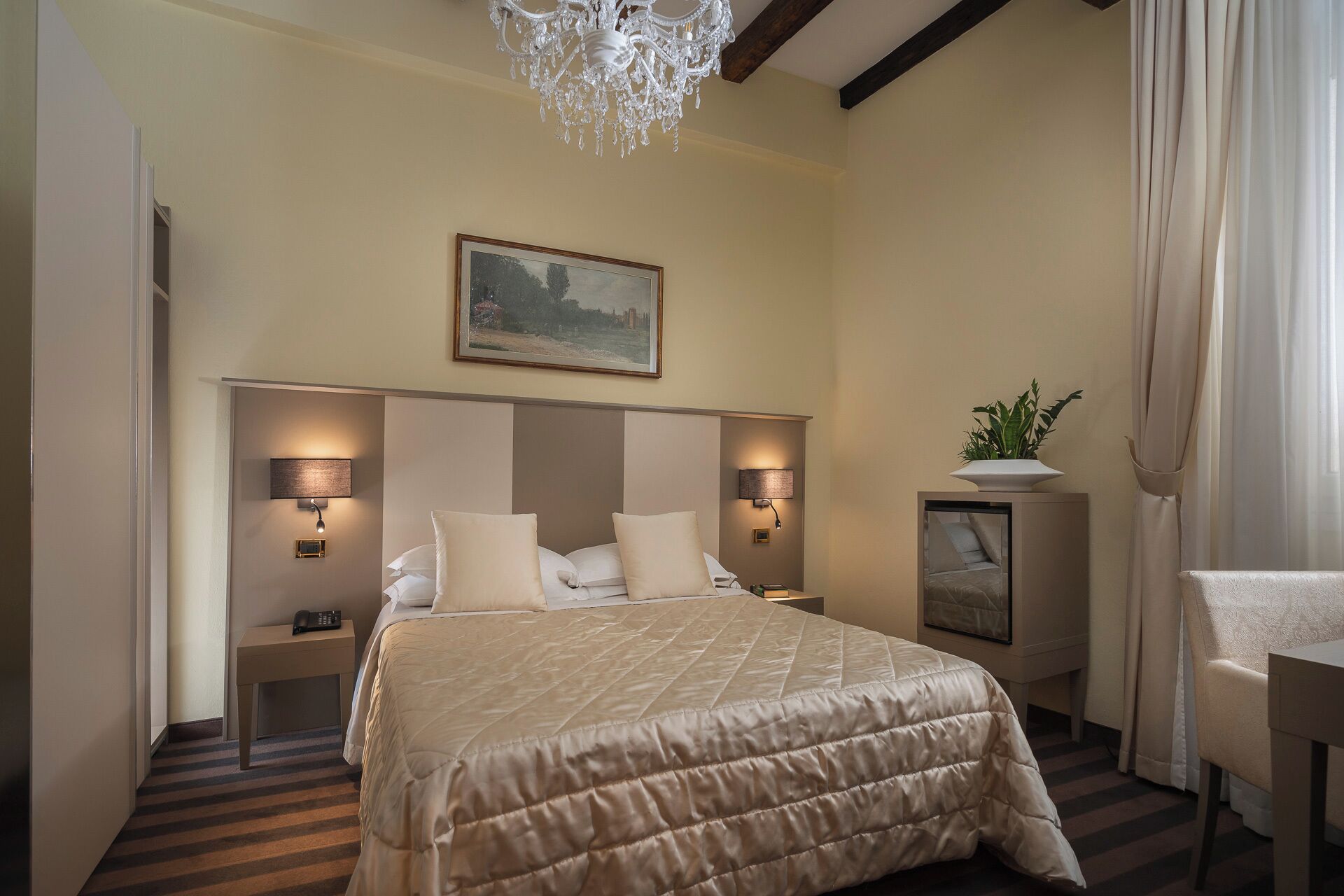 A hotel room with a large bed, two bedside lamps, a chandelier, a painting on the wall, a potted plant, and a chair.