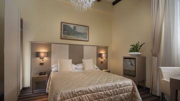 A hotel room with a large bed, two bedside lamps, a chandelier, a painting on the wall, a potted plant, and a chair.