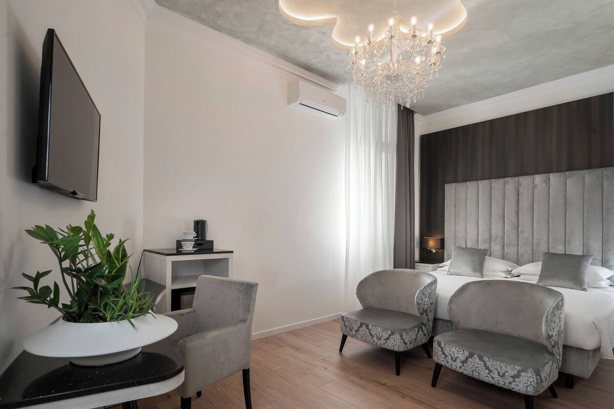 A modern hotel room with a large bed, two armchairs, a small table with a plant, a TV, and a chandelier.