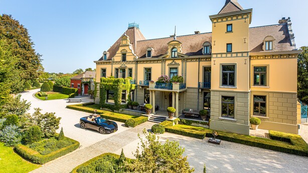Un grand bâtiment historique à étages, doté d’une tour imposante et d’un jardin bien entretenu, avec une allée et des voitures garées.