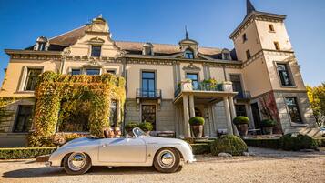 Une voiture décapotable blanche, un modèle classique, est garée devant un imposant bâtiment historique, doté d’une tour remarquable et d’une architecture richement décorée.