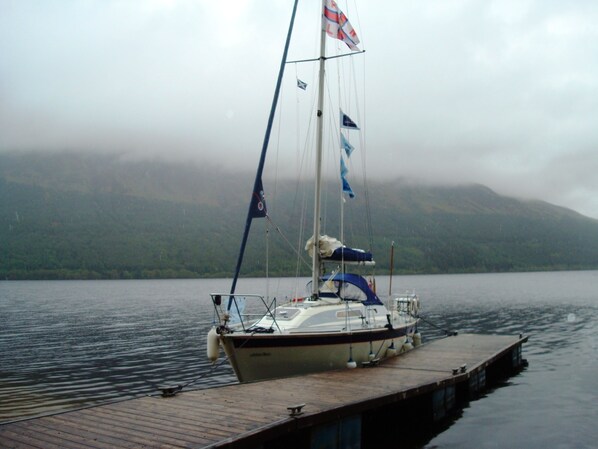 Een zeilboot afgemeerd aan een houten steiger, met een bergachtig landschap op de achtergrond.