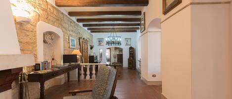 A spacious hallway with exposed wooden beams, a stone wall, and a fireplace. There is a wooden chair and a table with books and a lamp.