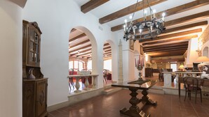 A spacious interior with a high ceiling, exposed wooden beams, and a central chandelier. There is a dining area with a table and chairs, and a kitchen area with a counter and chairs.