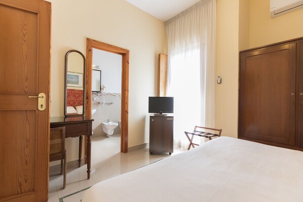 A hotel room with a bed, a television, a wooden wardrobe, a mirror, a bathroom visible through an open door, and a chair.
