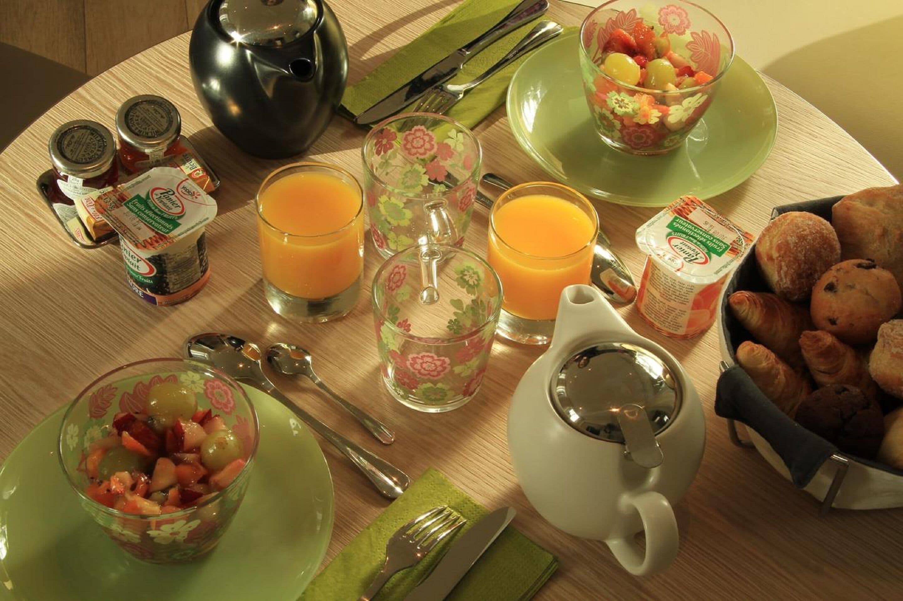 A breakfast setting with a teapot, cups, a bowl of fruit, and pastries on a wooden table.