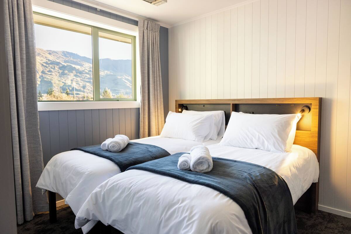 A double bed with white linens and a dark blue throw blanket, a wooden headboard, and a view of mountains through a window with curtains.