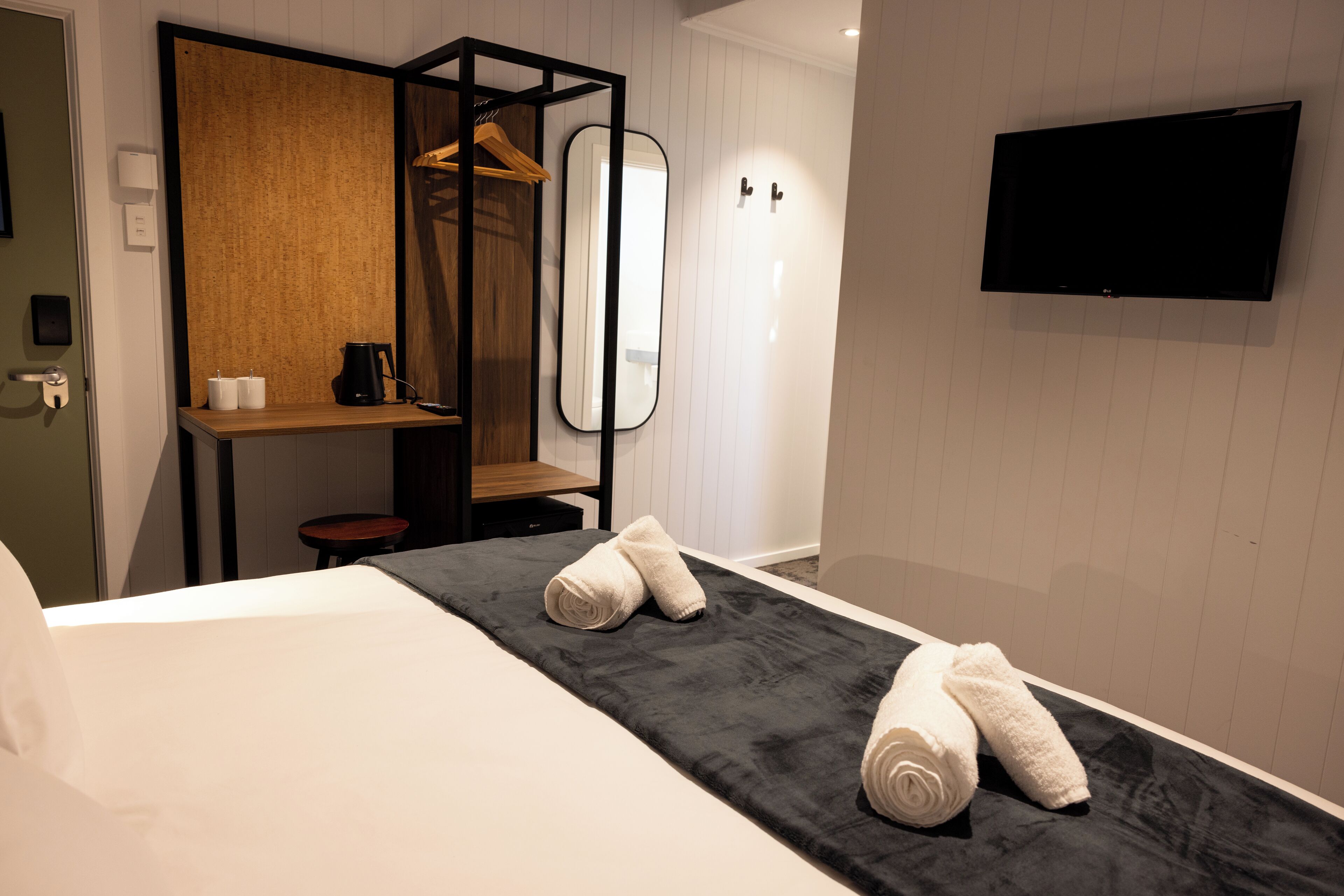 Habitación de hotel con cama, toalla negra, estante de madera, espejo y televisor de pantalla plana.