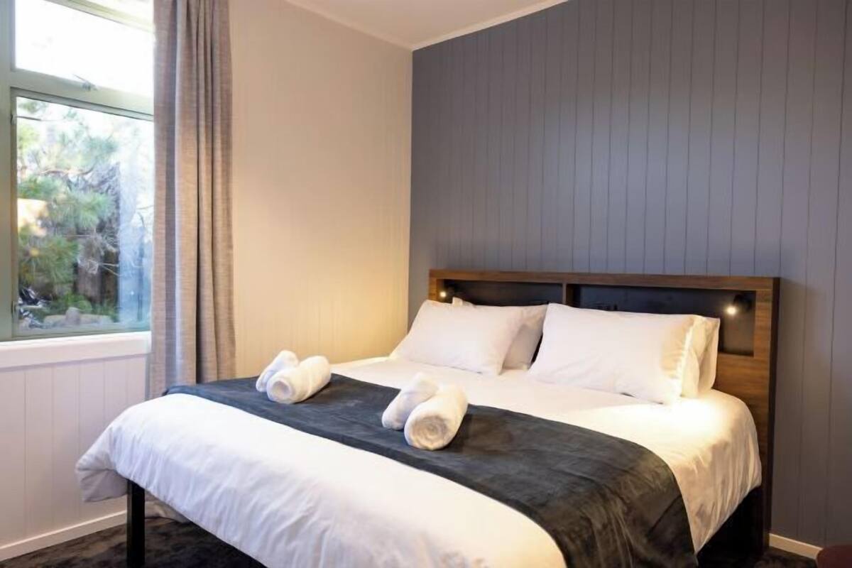 A neatly made bed with white linens and a dark headboard, a window with curtains, and a view of trees outside.