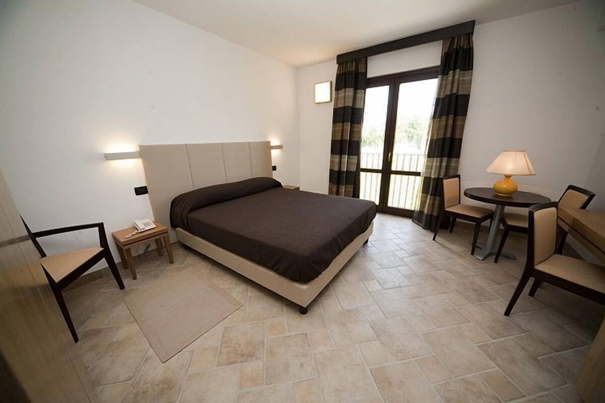 Ein Hotelzimmer mit Bett, Schreibtisch, Stuhl, einem kleinen Tisch mit Lampe und einem Fenster mit Vorhängen.