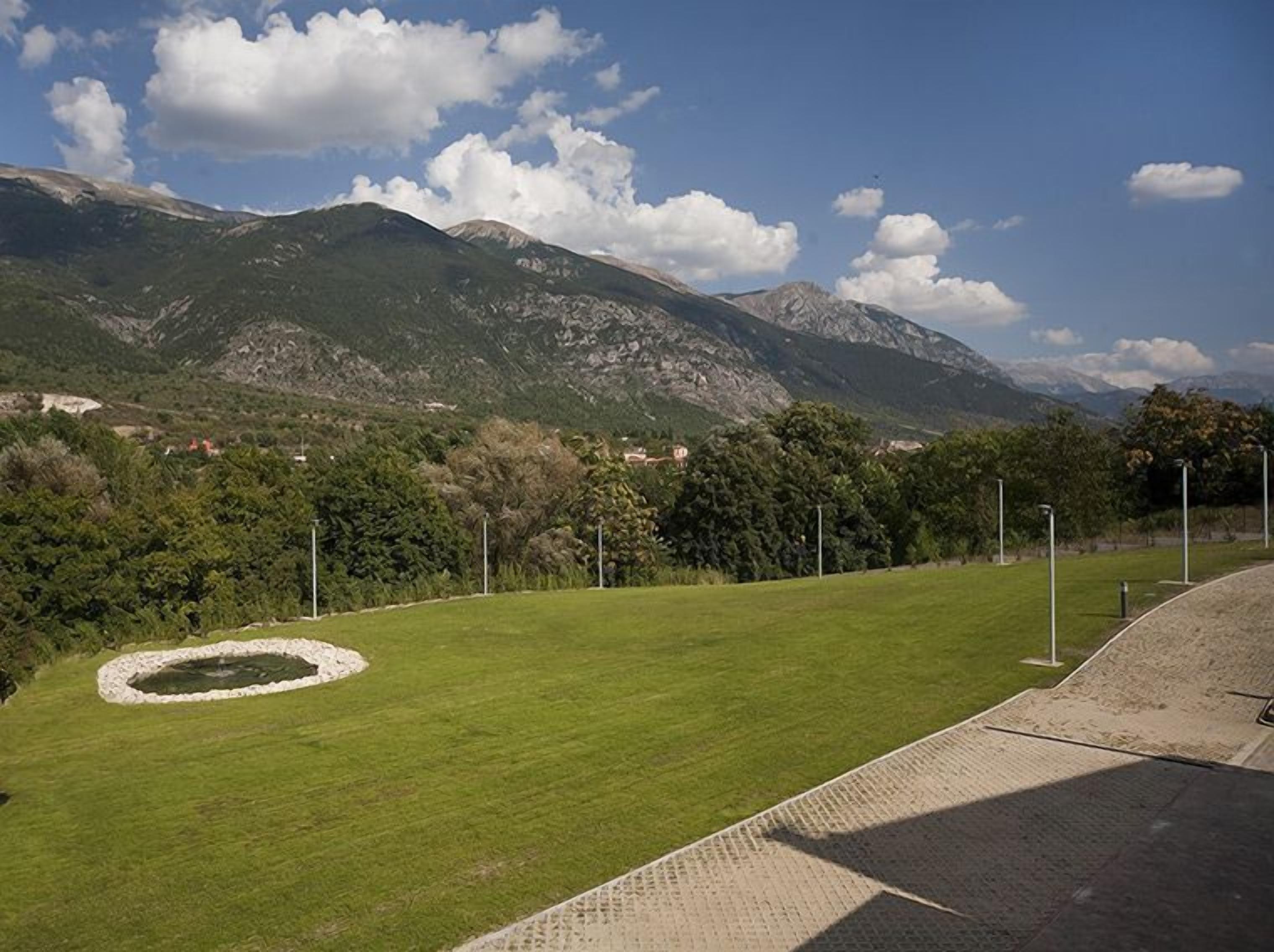 A grassy field with a circular stone structure, surrounded by trees and mountains in the background.