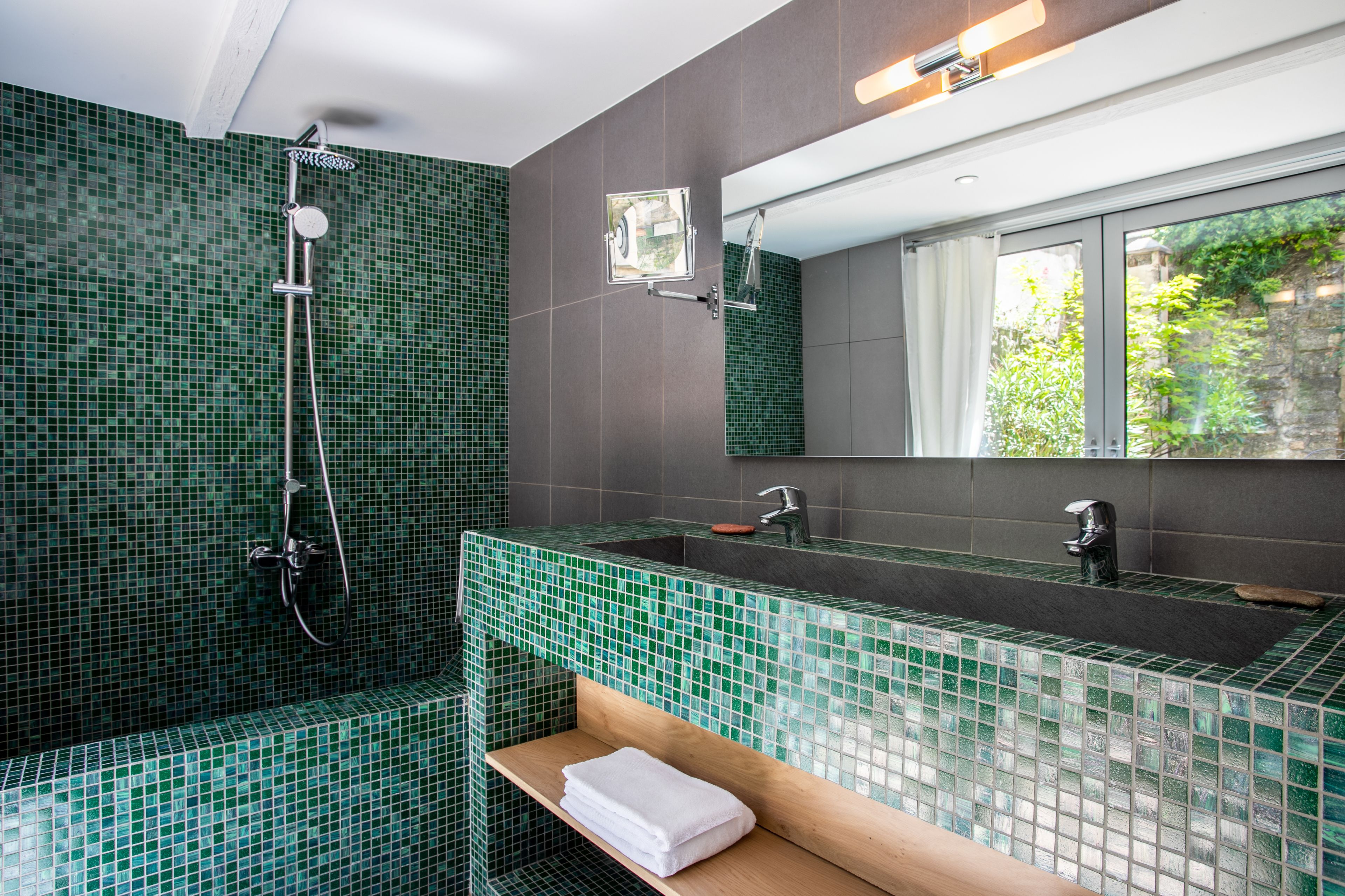A bathroom with a green mosaic tile shower, a large mirror, and a window providing a view of greenery outside.