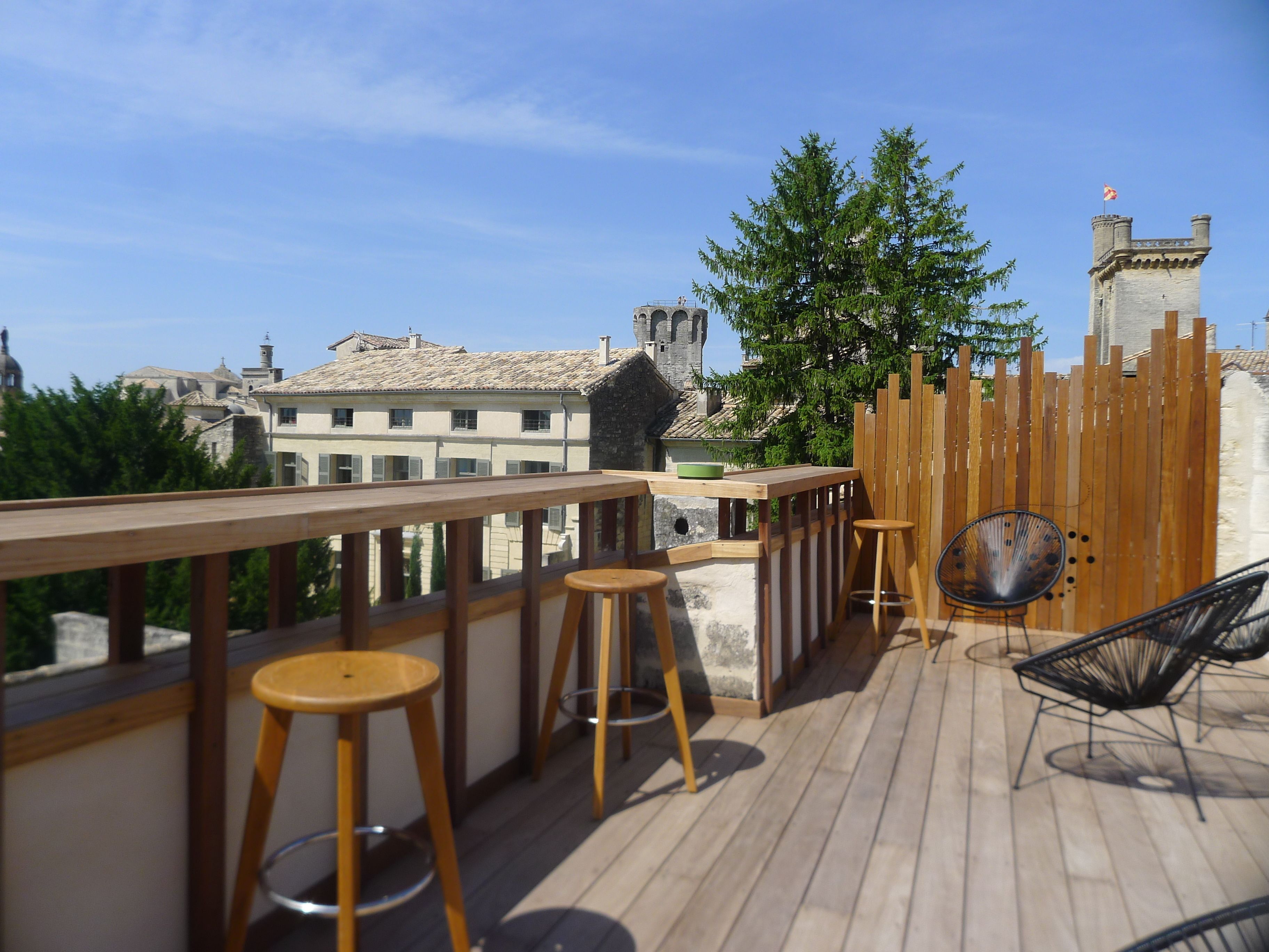 Una terraza de madera con taburetes de bar y una silla, con vistas a un paisaje urbano que incluye edificios históricos y una bandera en una torre.