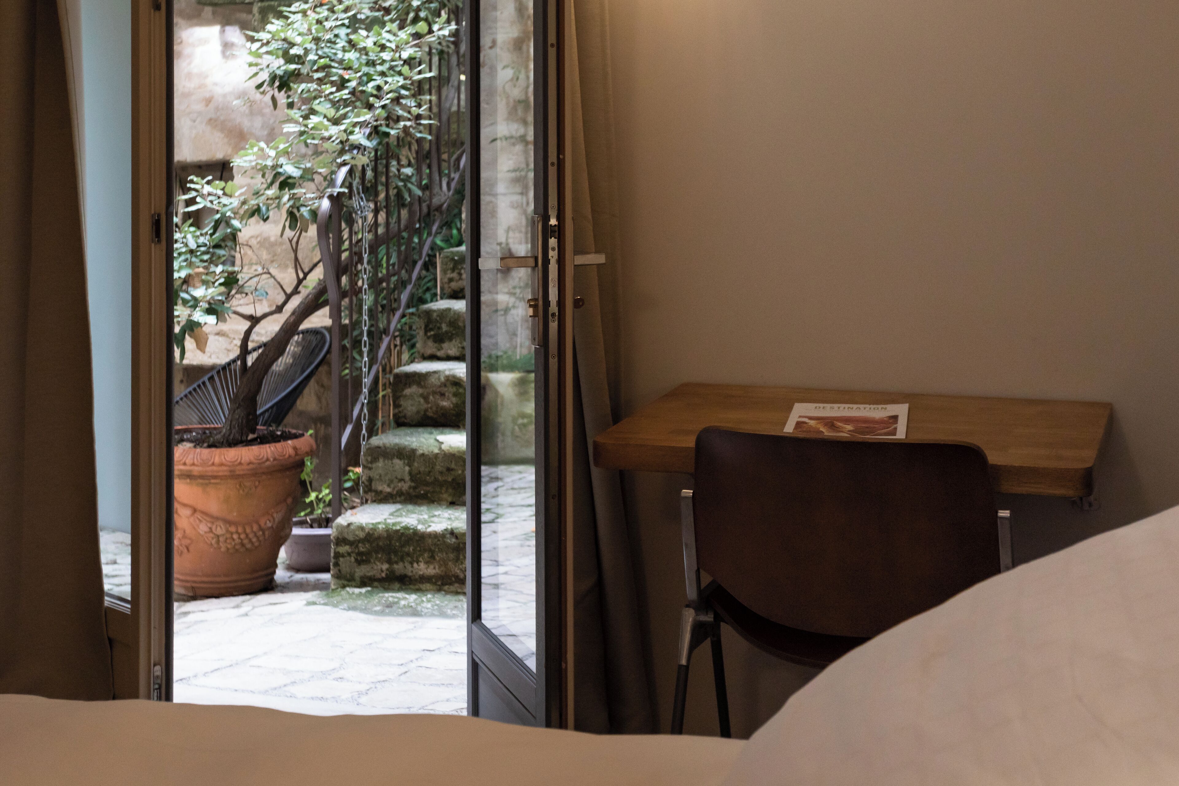 Una habitación con un escritorio de madera, una silla y una cama. Se ve una ventana con vistas a un jardín y una escalera que da al exterior.