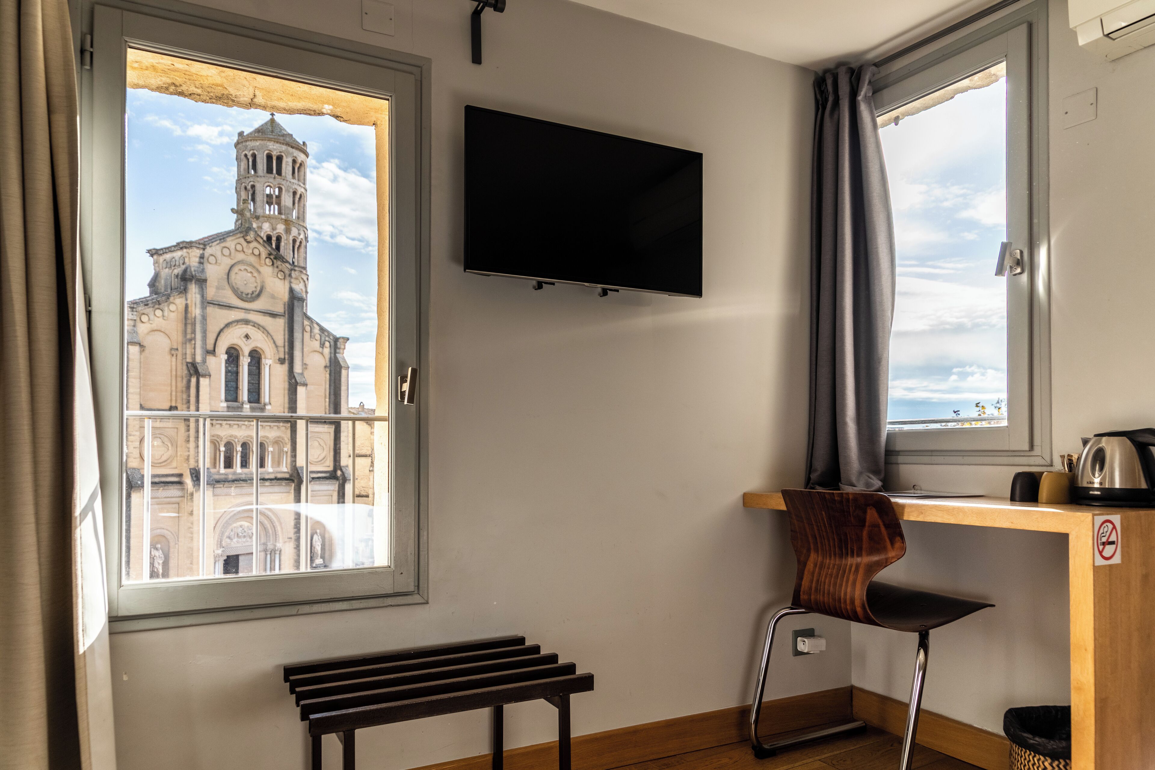 Une pièce dotée d’une grande fenêtre donnant sur un bâtiment historique, d’un téléviseur à écran plat fixé au mur, d’un bureau en bois, d’une chaise et d’un banc.