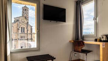 Une pièce dotée d’une grande fenêtre donnant sur un bâtiment historique, d’un téléviseur à écran plat fixé au mur, d’un bureau en bois, d’une chaise et d’un banc.