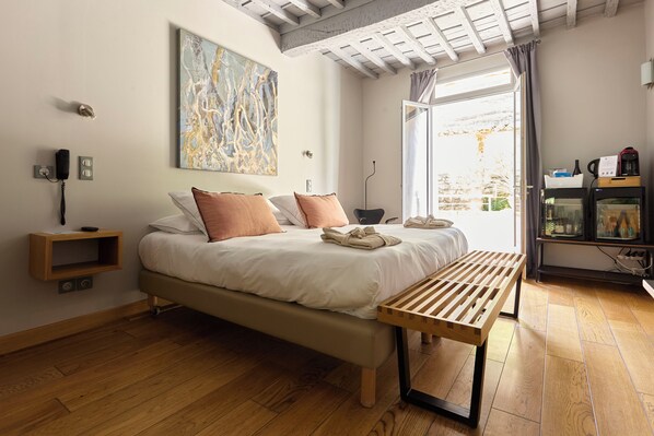Un dormitorio con cama, mesita de noche, banco, cuadro, teléfono y ventana con cortinas.