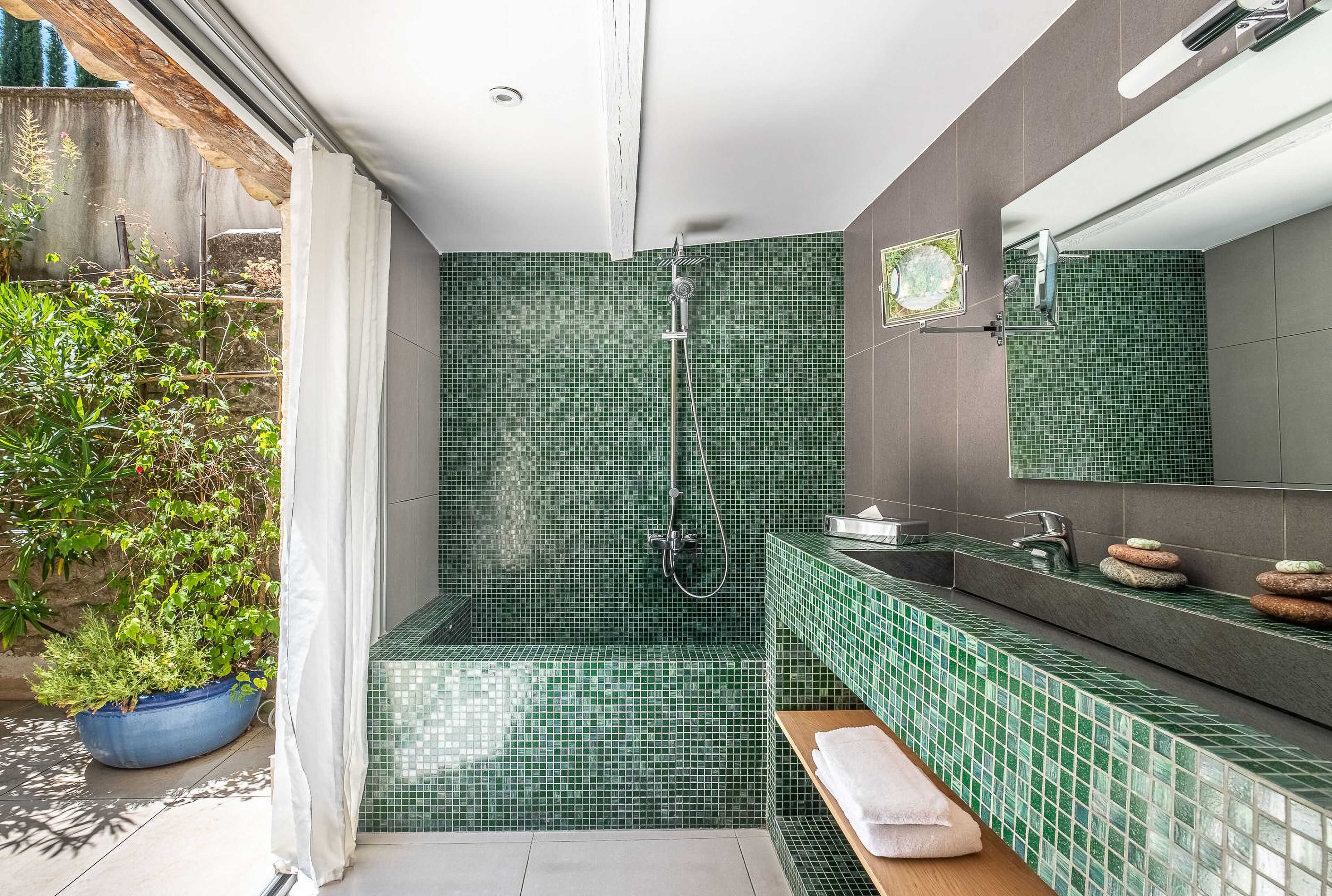 A bathroom with green mosaic tiles, a shower, a sink with a mirror, and a potted plant.