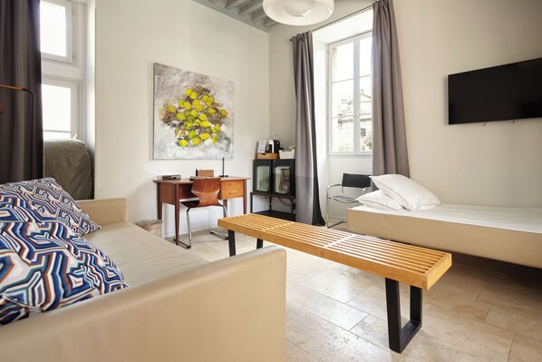 Un dormitorio moderno con cama, banca, escritorio, silla, televisor y ventana con cortinas.