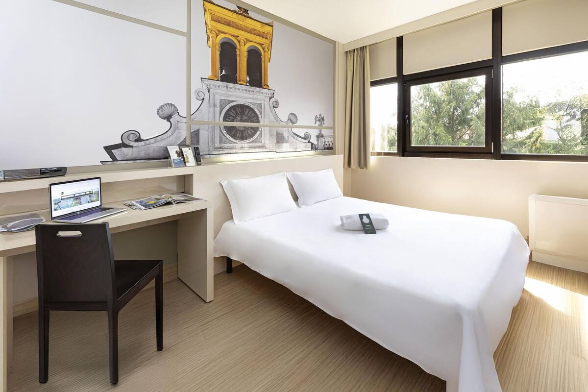 Une chambre d’hôtel avec un lit, un bureau, une chaise et une grande fenêtre.