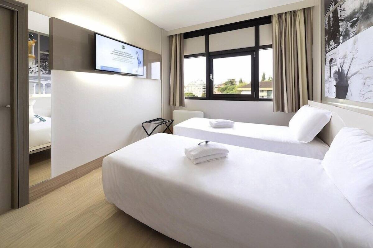 Une chambre d’hôtel avec un lit, un bureau, une chaise, une télévision et une fenêtre avec des rideaux.