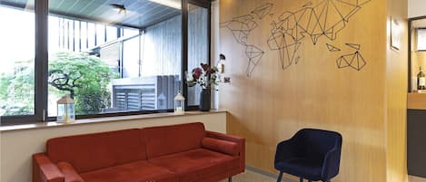 Un área de recepción moderna con un sofá rojo, una silla azul oscuro, una pared de madera con arte abstracto, una ventana con vistas a la vegetación y una mesa pequeña con revistas y una planta.