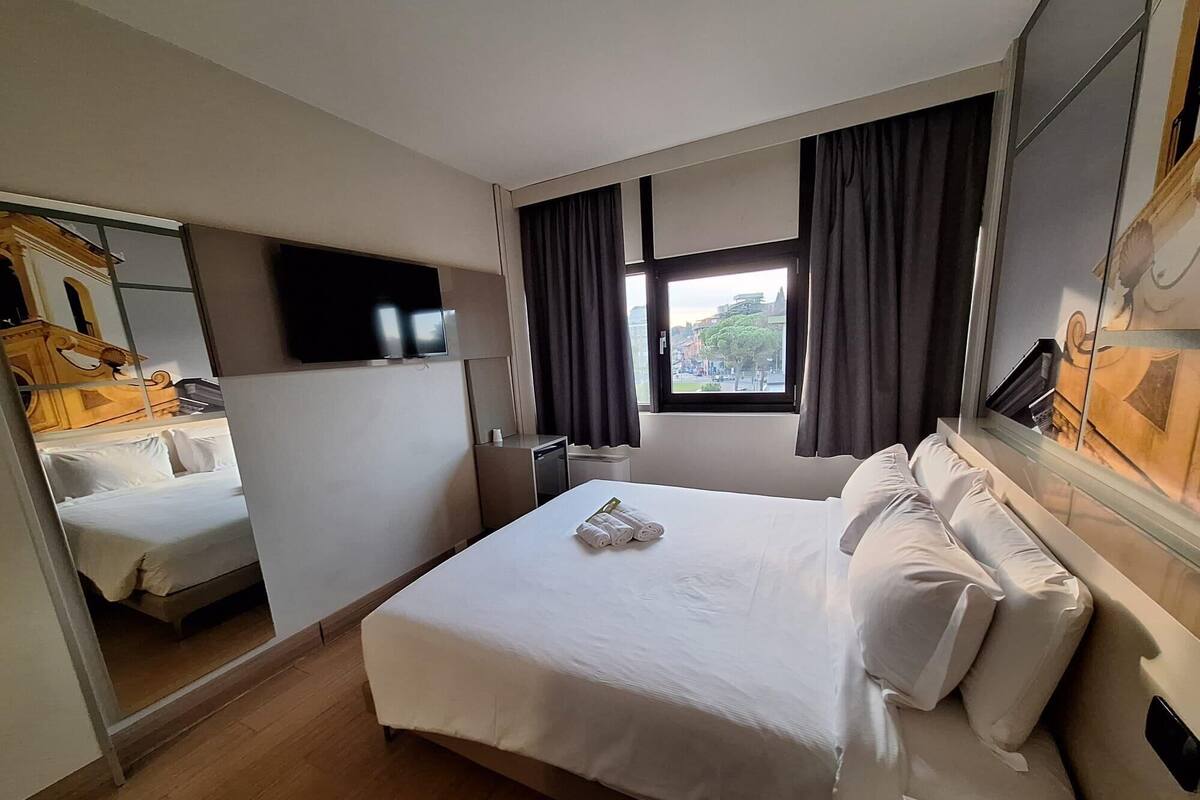 Une chambre d’hôtel avec un grand lit, un téléviseur fixé au mur, une fenêtre avec des rideaux et un plancher en bois.