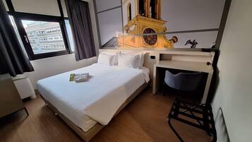 Quarto de hotel com cama, escrivaninha, cadeira, relógio e janela com cortinas.
