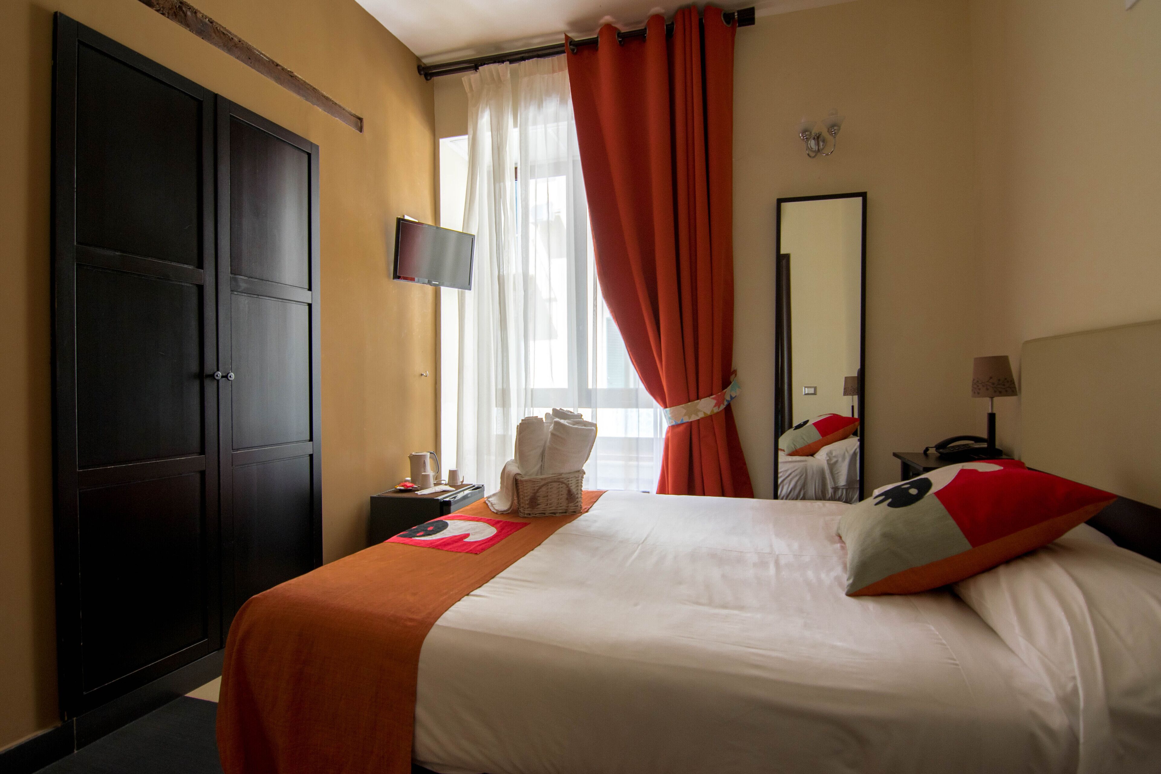 A hotel room with a large wardrobe, a bed with white and orange bedding, a flat-screen TV, a mirror, and a window with red curtains.