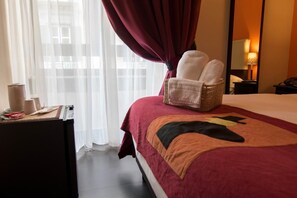 A hotel room with a bed, a chair with towels, a desk with a lamp, and a window with curtains. - Hotel Plebiscito Aparthotel (Naples)