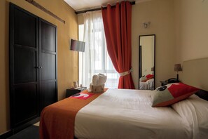A hotel room with a large wardrobe, a bed with white and orange bedding, a flat-screen TV, a mirror, and a window with red curtains. - Hotel Plebiscito Aparthotel (Naples)