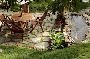Un patio de piedra con una mesa redonda de madera y sillas, rodeado por un pequeño estanque y abundante vegetación.