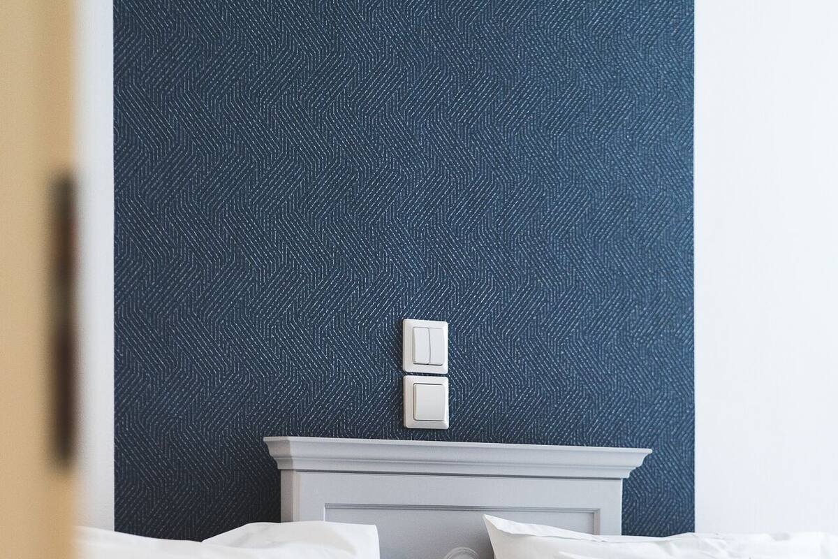 A neatly made bed with white linens and a grey throw blanket, a white nightstand with a switch, and a dark blue textured wall.