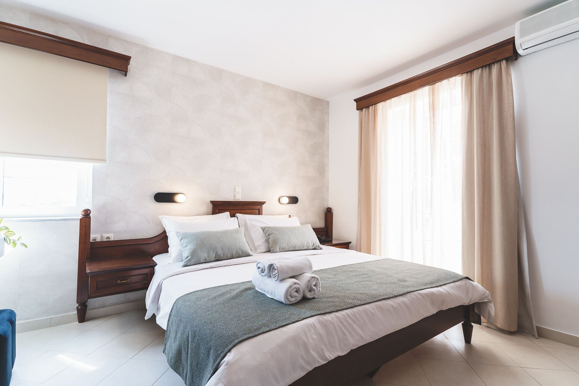 Ein ordentlich bezogenes Bett mit grauer Decke und gerollten Handtüchern, ein Holz-Kopfstueck, Nachttische und ein Fenster mit Vorhängen.