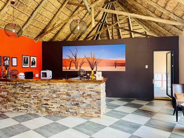 A reception area with a thatched roof, a checkered floor, and a large painting of a desert landscape. - Agama Lodge (Sesriem)