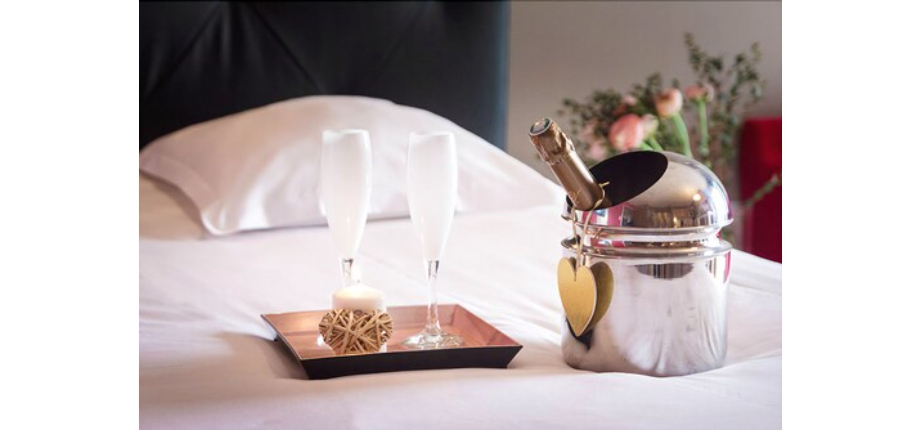 Ein ordentlich angerichteter Tablett mit zwei Sektgläsern, einer Kerze und einem dekorativen Gegenstand auf einem Bett, im Hintergrund ein Kissen und eine Blumenvase.
