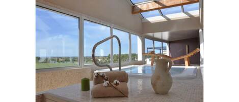 Un intérieur moderne avec de grandes fenêtres, une structure métallique incurvée et un petit canapé beige agrémenté d’un vase.