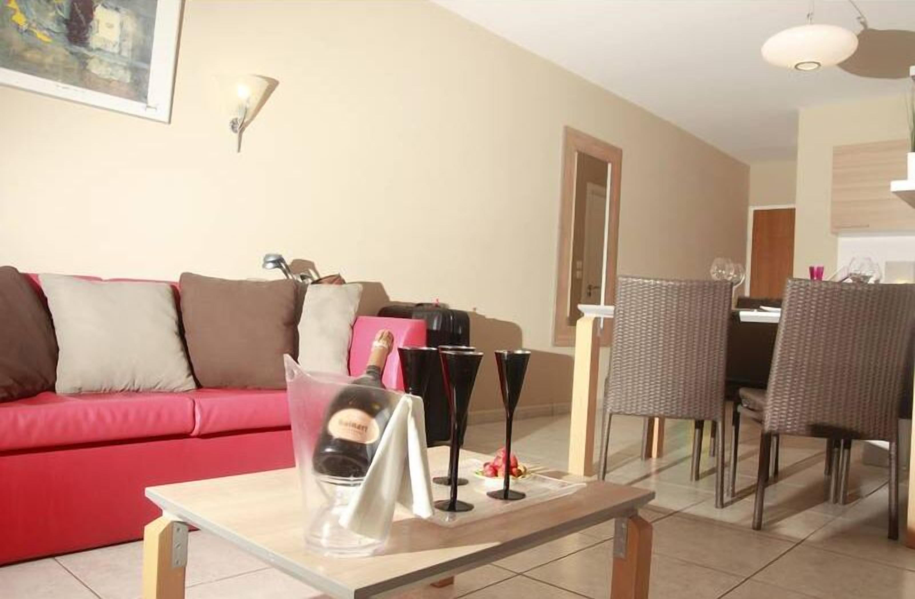 Uma sala de estar com um sofá vermelho, uma área de jantar com cadeiras de vime e uma mesa com garrafa e copos.