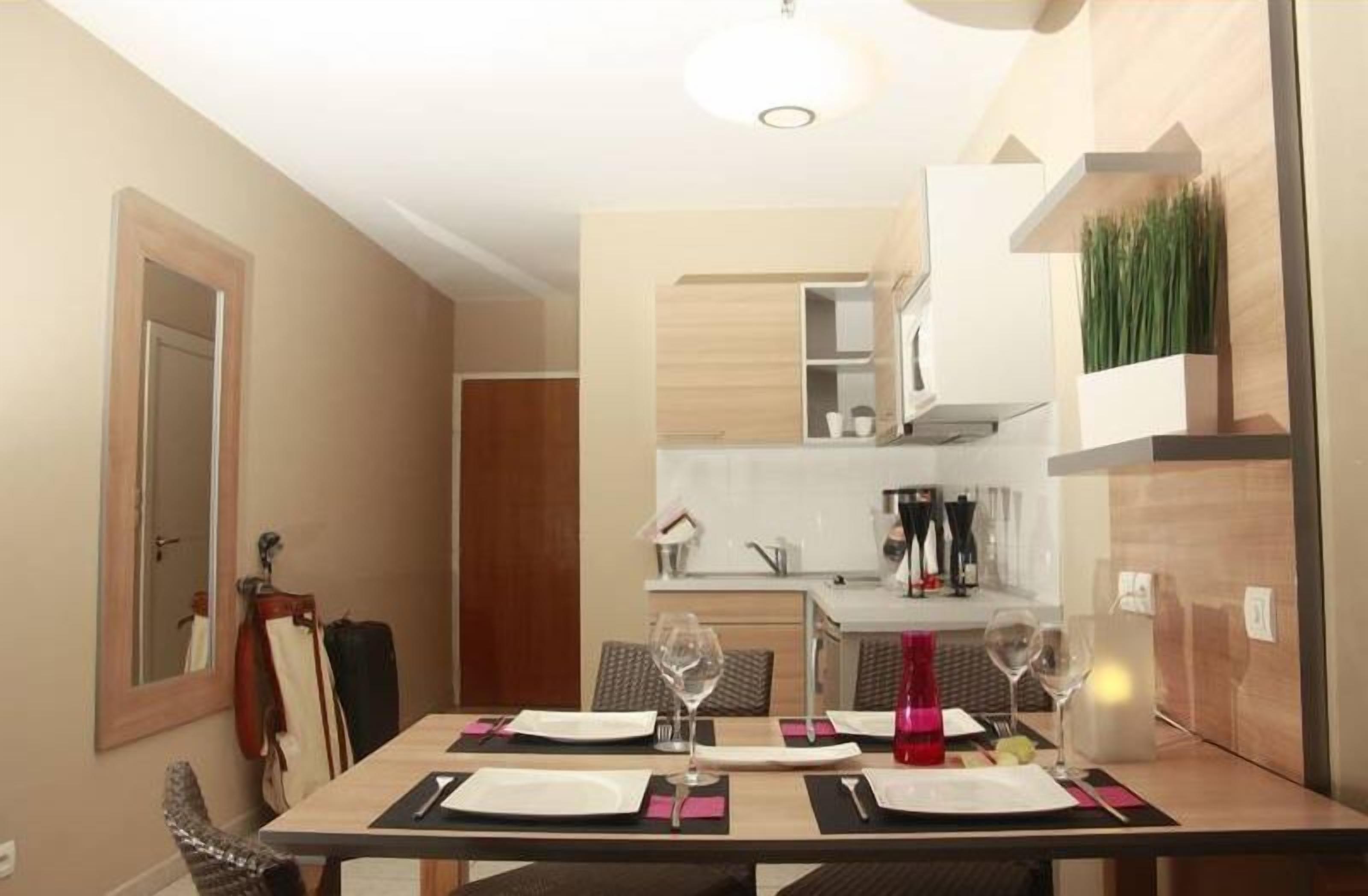 Un coin repas avec une table dressée pour quatre personnes, une cuisine en arrière-plan et une porte sur la gauche.