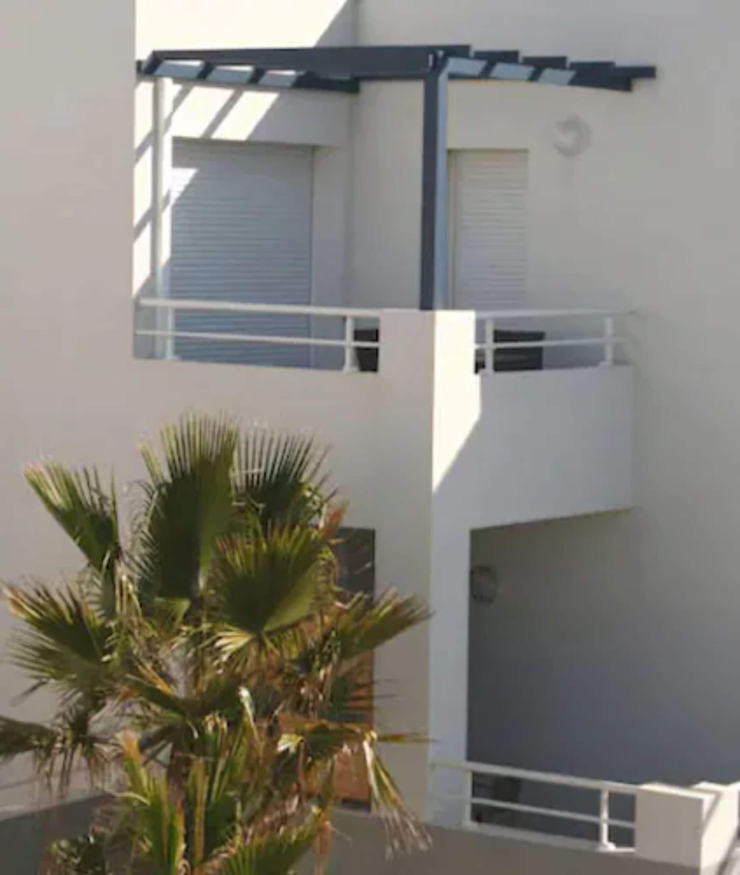 A modern two-story building with balconies and a palm tree in the foreground.