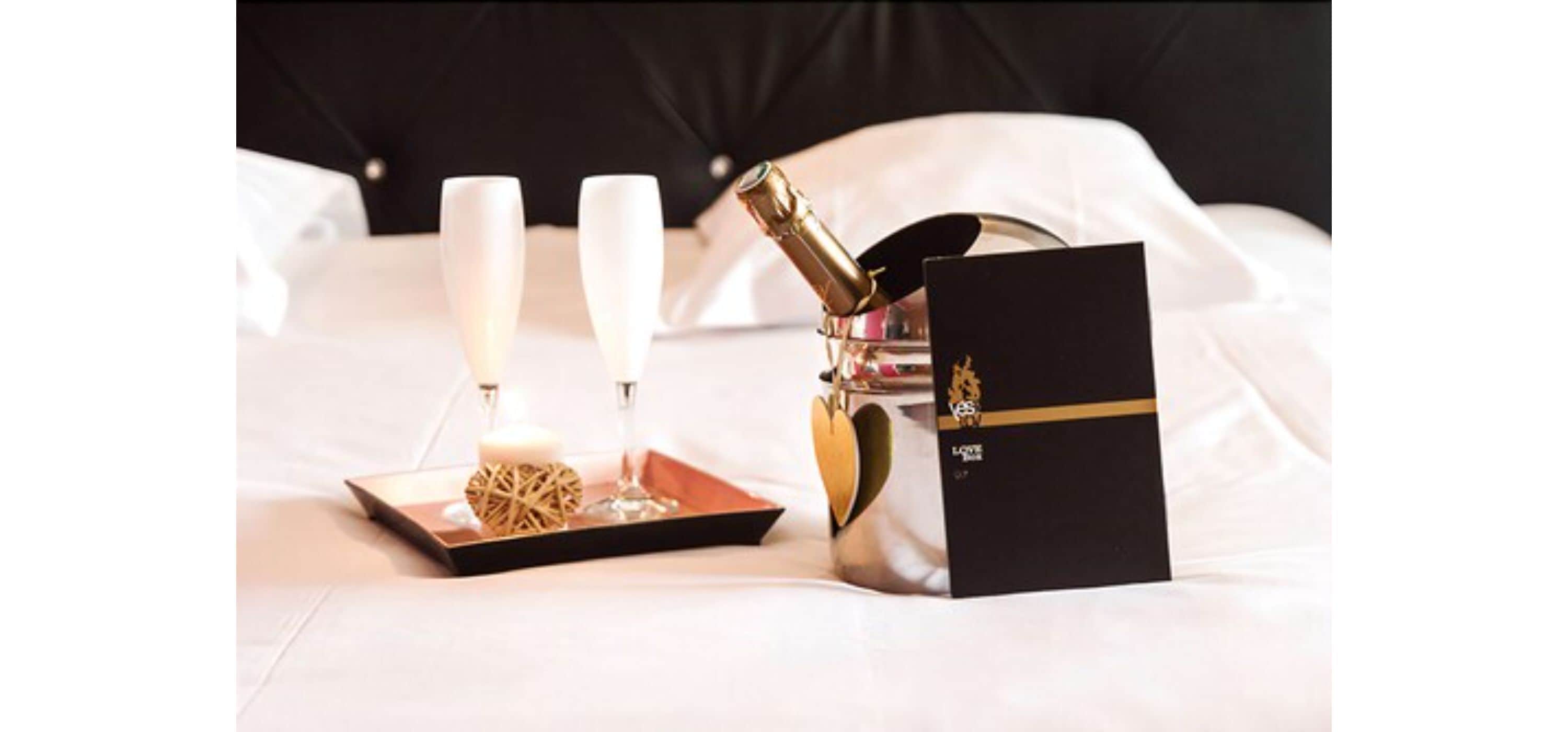 A bed with white linens, a champagne bottle, two glasses, and a black box with gold lettering.