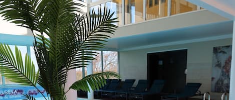 An indoor swimming pool with a wooden deck, lounge chairs, and a large potted plant.