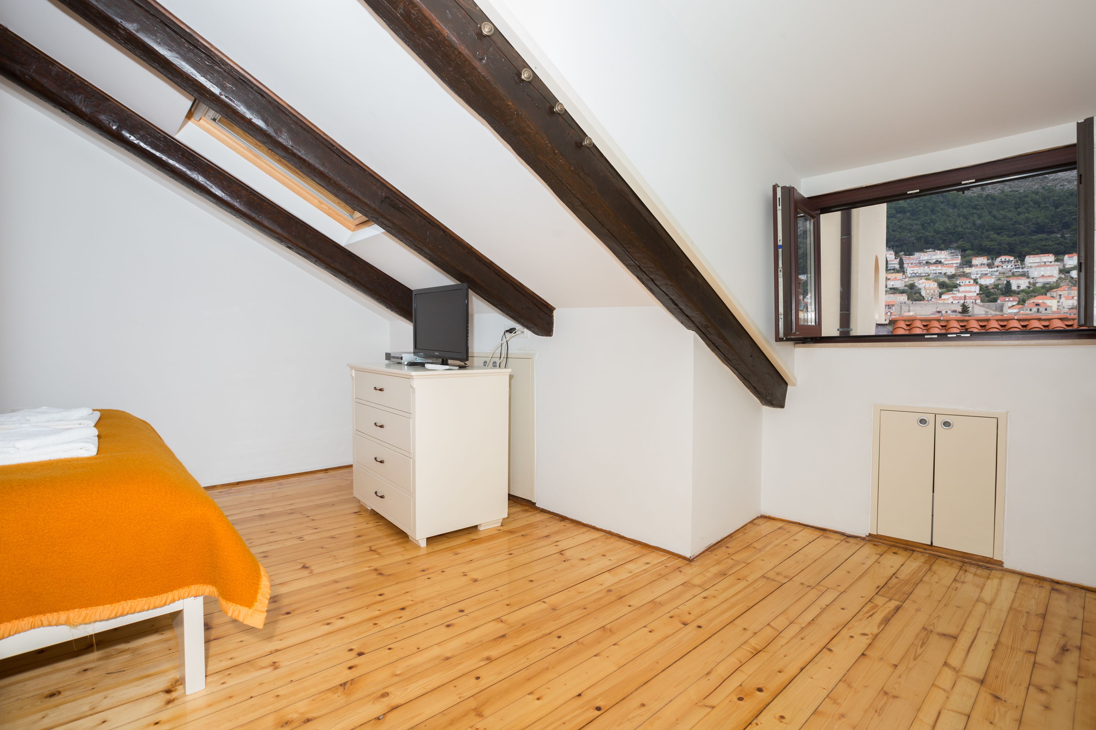 A bedroom with a sloped ceiling, a bed with an orange blanket, a dresser, and a window with a view of a town.