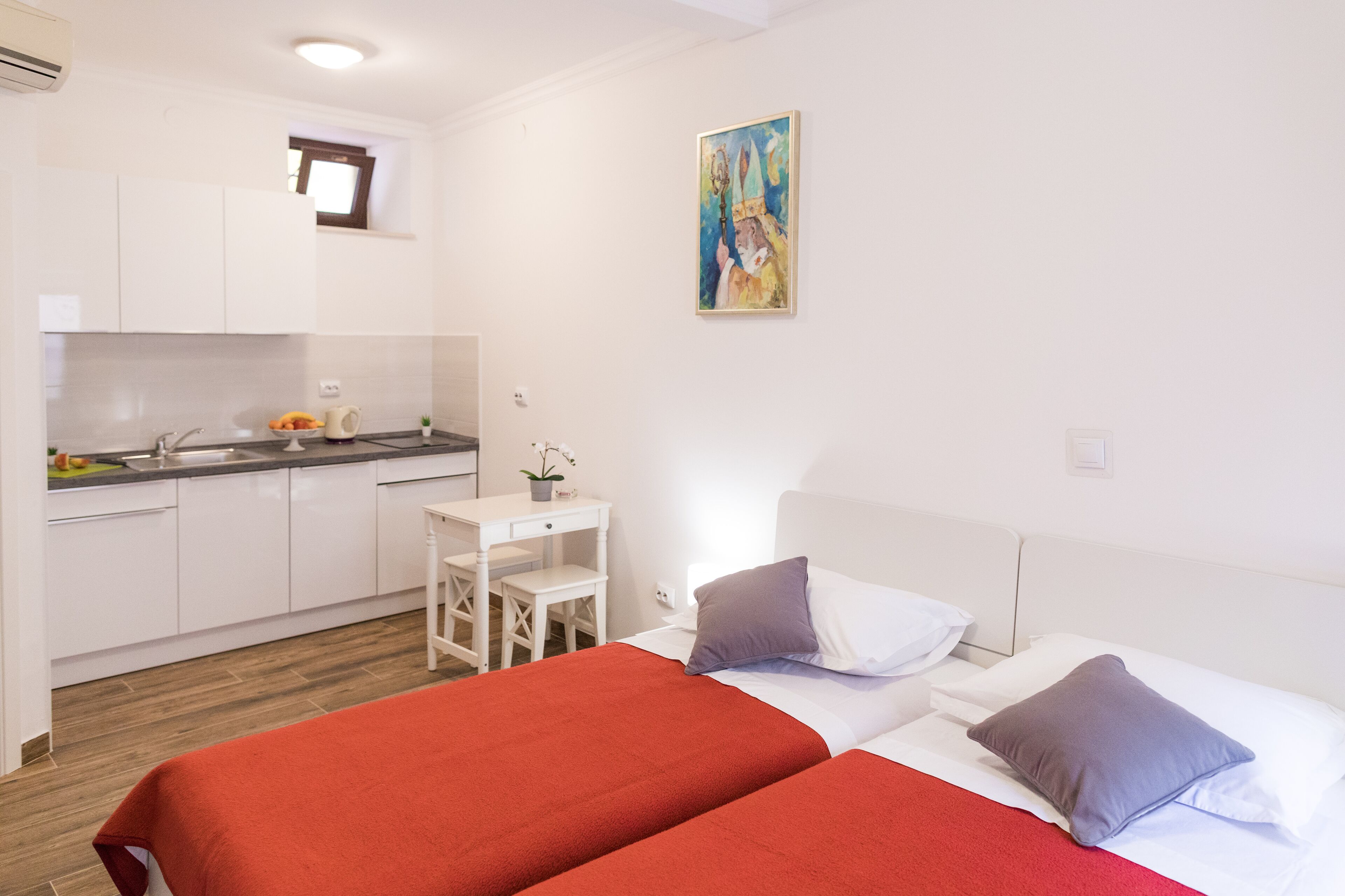 A hotel room with two beds, a small desk, a kitchenette, and a painting on the wall.