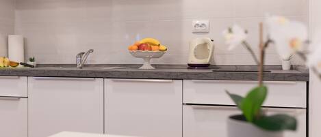 Una cocina moderna con armarios blancos, encimera de mármol, frutero, hervidor y planta en maceta.