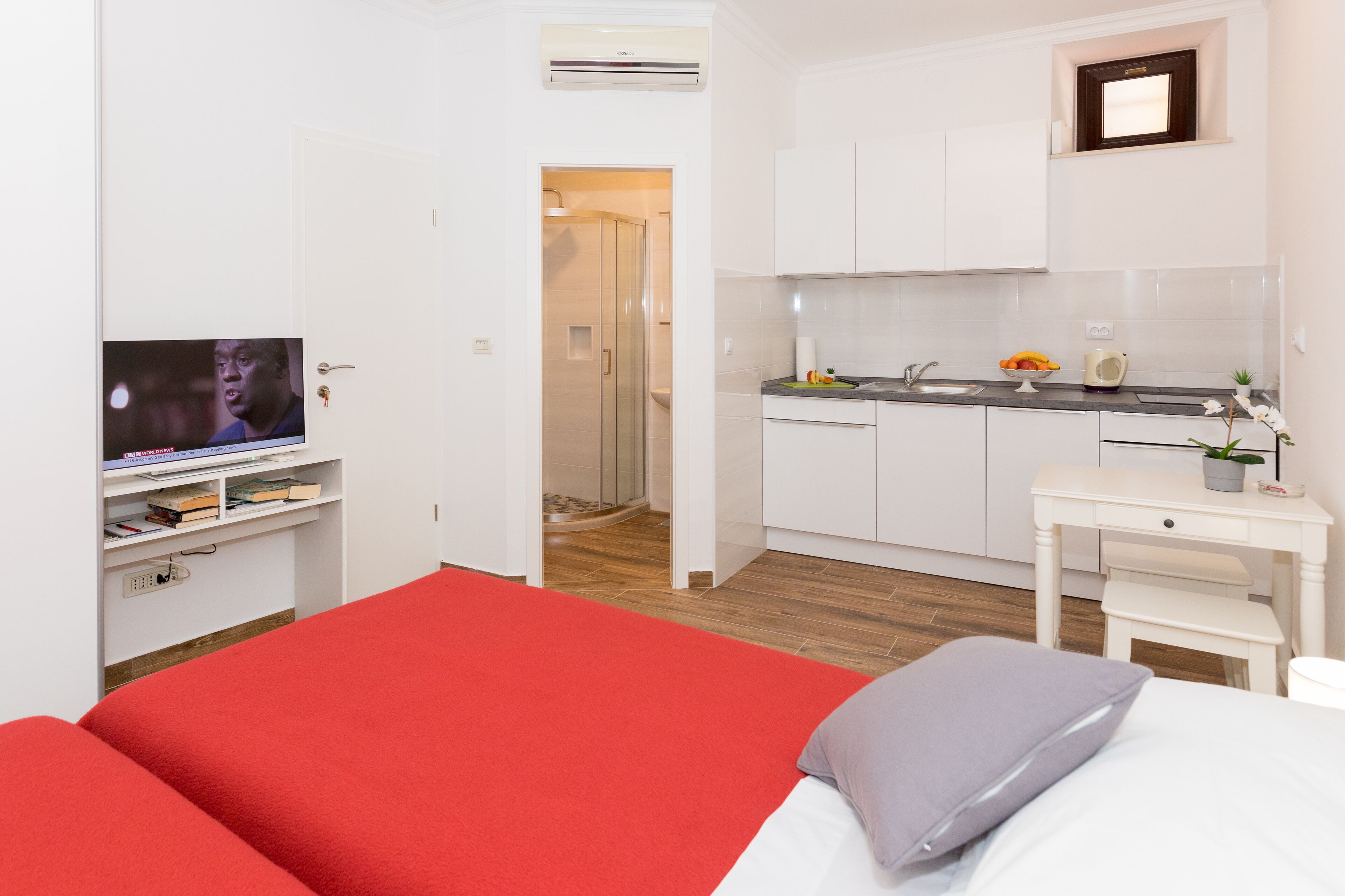 Een hotelkamer met een rood bed, een klein wit bureau, een kitchenette en een televisie op een standaard.