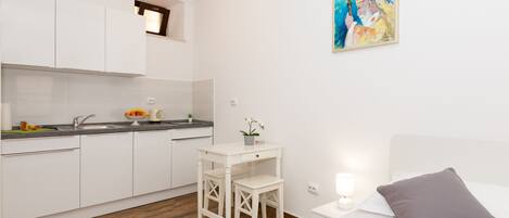 A compact hotel room with a kitchenette, a small dining table, a bed with a red blanket, and a painting on the wall.