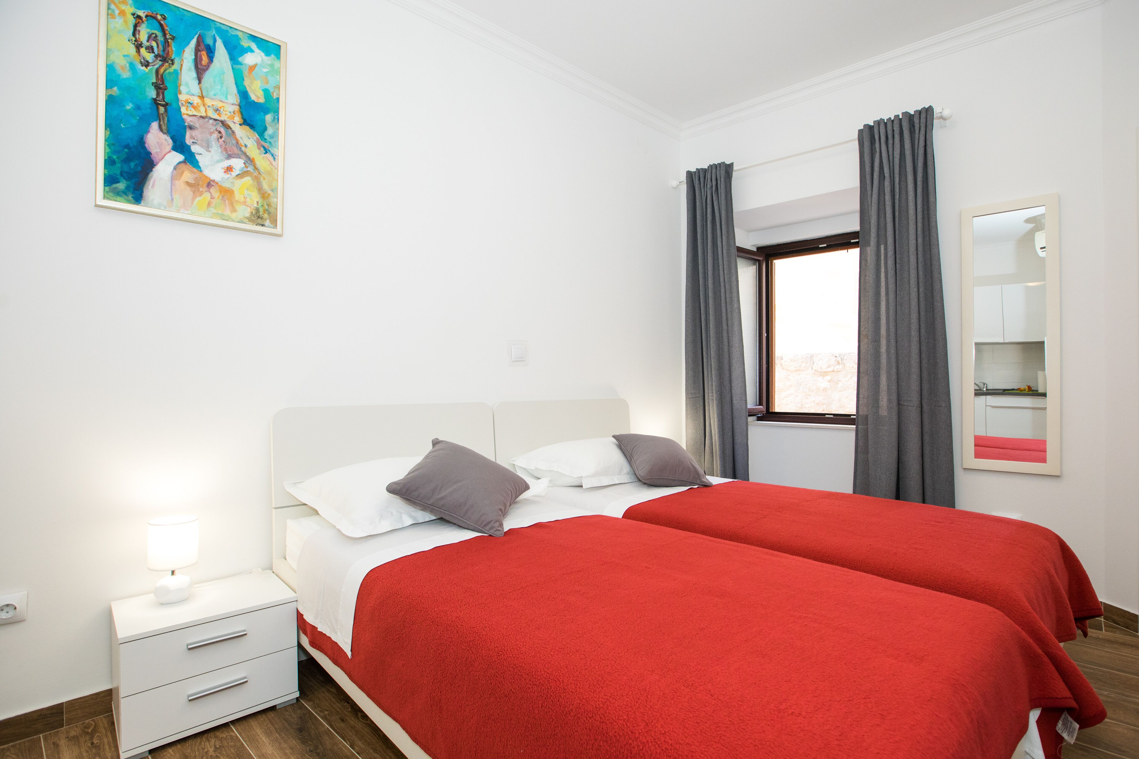 A hotel room with two beds, a nightstand, a lamp, a painting, a window with curtains, and a mirror.
