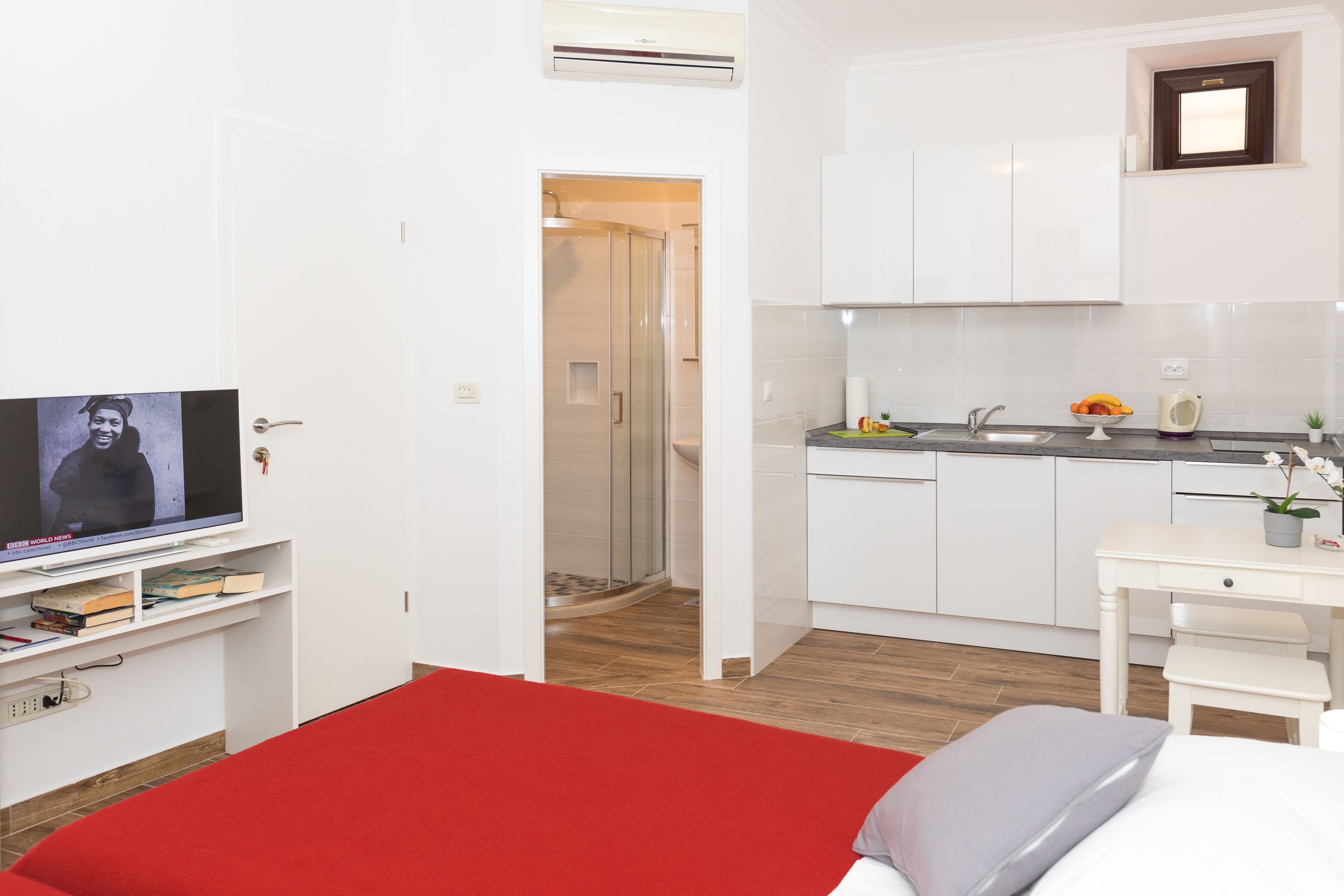 A modern hotel room with a red bed, a TV, a kitchenette, and a shower.