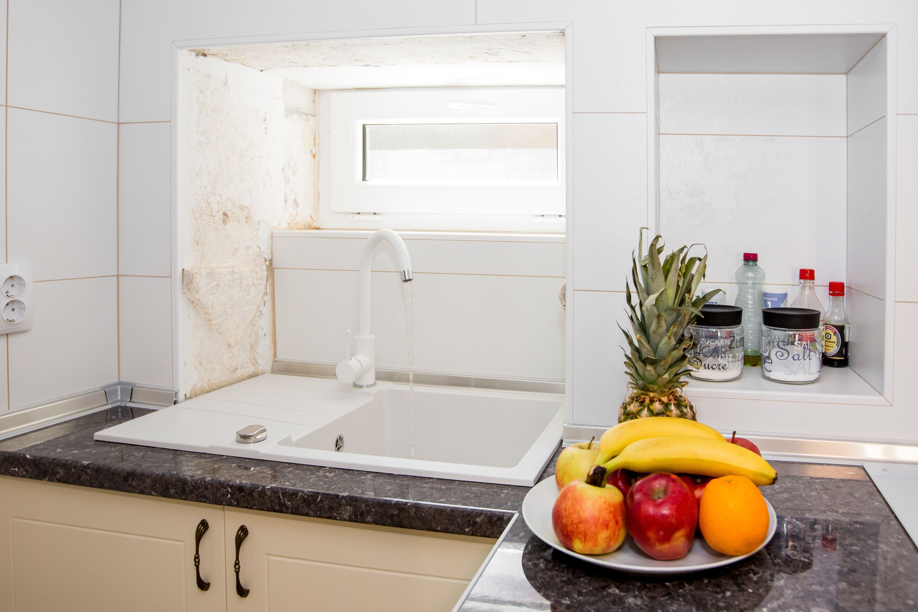 A kitchen with a white sink, a granite countertop, and a fruit bowl containing apples, bananas, and an orange.